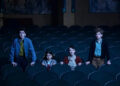 Four children stand among empty theater seats, looking intently ahead, suggesting a scene from a movie or show