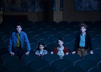 Four children stand among empty theater seats, looking intently ahead, suggesting a scene from a movie or show