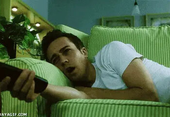 Person lying on a couch, holding a TV remote, with a bored expression while watching television