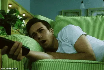 Person lying on a couch, holding a TV remote, with a bored expression while watching television