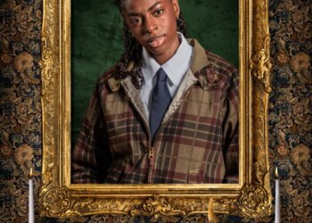 Singer Cat Burns, a contestant on "The Celebrity Traitors," dressed in a plaid jacket, white shirt, and navy tie, framed by a gold ornate picture frame with a floral tapestry background.