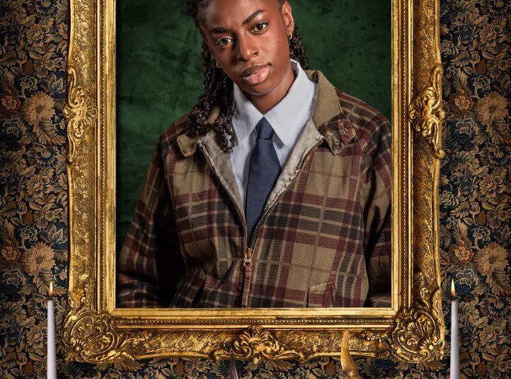 Singer Cat Burns, a contestant on "The Celebrity Traitors," dressed in a plaid jacket, white shirt, and navy tie, framed by a gold ornate picture frame with a floral tapestry background.