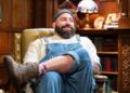 Man with large beard and wearing beanie sits with one leg crossed over the other and smiles at someone not pictured.