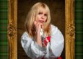 Woman with blonde hair wearing puffy white shirt stares into the camera with her head resting on her fist.