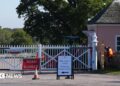 A white gate, Cranbourne Gate, in Windsor Great Park, with two workmen stood to the right of it. One is wearing an orange jacket and the other a green jacket. There is a red "road closed" sign in front of the gate and a sign to its right which says: "This gate is closed, please use Rangers Gate".