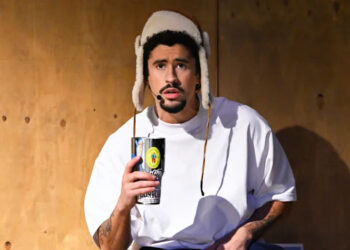 Bad Bunny wearing a white shirt and a furry hat holds a can, sitting against a wooden wall.
