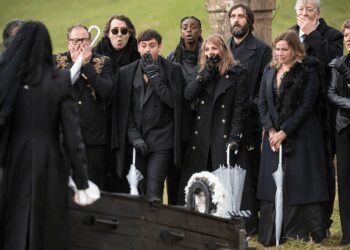 A group of funeral mourners gasp in front of an open coffin