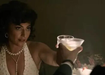 A person in a glamorous, vintage-style outfit shares a toast at an elegant setting, holding a drink and a cigarette, with flowers in the background