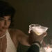 A person in a glamorous, vintage-style outfit shares a toast at an elegant setting, holding a drink and a cigarette, with flowers in the background
