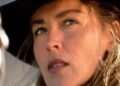 A close-up of a woman with light brown hair wearing a wide-brimmed black hat, looking thoughtfully to the side. Another blurred figure is partially visible in the foreground.