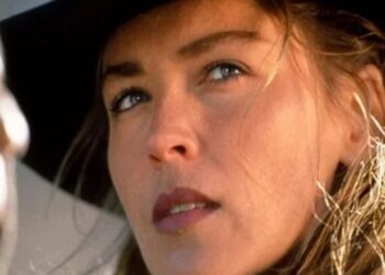 A close-up of a woman with light brown hair wearing a wide-brimmed black hat, looking thoughtfully to the side. Another blurred figure is partially visible in the foreground.