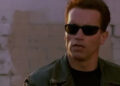 Person in a leather jacket and sunglasses looking to the side with a serious expression, against a brick wall background