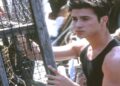 A young man with short dark hair wearing a sleeveless black shirt stands outdoors, holding onto a wooden structure with wire mesh, possibly a lobster trap, while looking off to the side.
