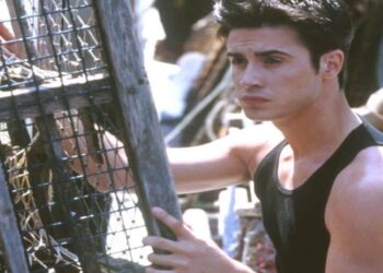 A young man with short dark hair wearing a sleeveless black shirt stands outdoors, holding onto a wooden structure with wire mesh, possibly a lobster trap, while looking off to the side.