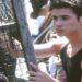 A young man with short dark hair wearing a sleeveless black shirt stands outdoors, holding onto a wooden structure with wire mesh, possibly a lobster trap, while looking off to the side.
