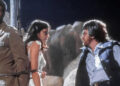 A man and woman are tied together with rope while another man faces them, speaking seriously. They are outdoors, surrounded by large rocks, and it appears to be nighttime.
