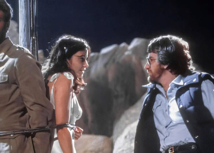 A man and woman are tied together with rope while another man faces them, speaking seriously. They are outdoors, surrounded by large rocks, and it appears to be nighttime.