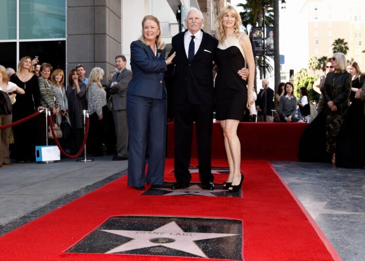 Three people in formal attire stand on a red carpet with a large star embedded in the ground.