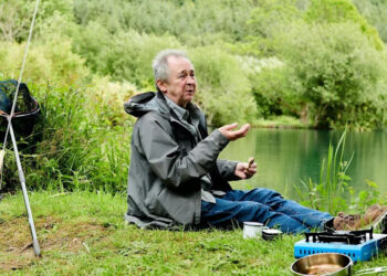 Paul sitting on the grass next to a green lake. There is a fish net and rod in a green plant bush. He is holding food in his hand. On the ground, there is a blue travel gas cooker, bronze coloured pot and a white cup near Paul's brown boots.