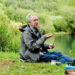 Paul sitting on the grass next to a green lake. There is a fish net and rod in a green plant bush. He is holding food in his hand. On the ground, there is a blue travel gas cooker, bronze coloured pot and a white cup near Paul's brown boots.