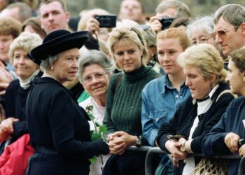 Queen Elizabeth, in a black coat and hat, wears a worried expression on her face while greeting a crowd of women