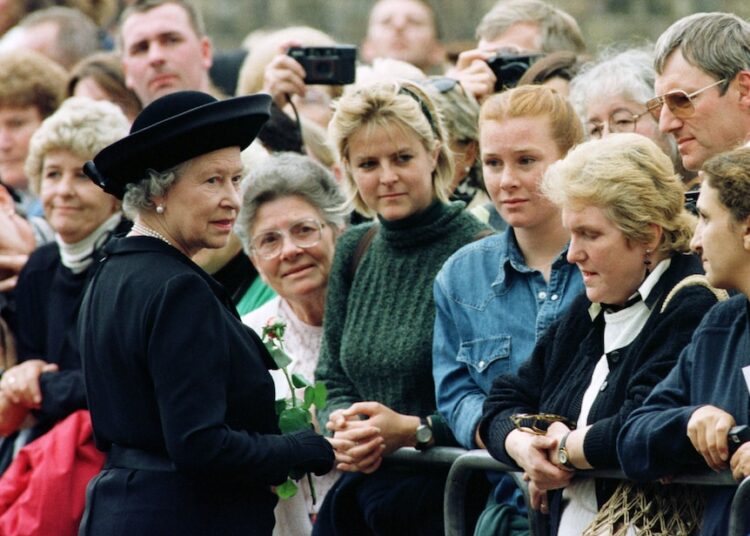 Queen Elizabeth, in a black coat and hat, wears a worried expression on her face while greeting a crowd of women