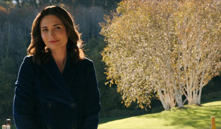 Zosia smiling slightly standing in front of a tree on Pluribus