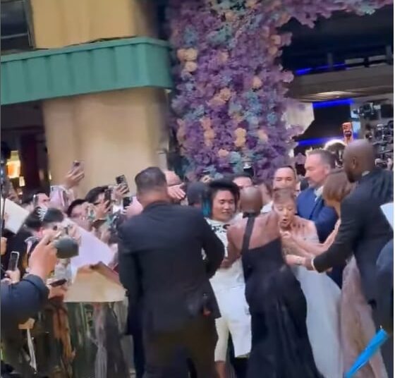 Fans line up behind a barrier. On a bright yellow carpet, security move toward women in gowns to remove a man in a tshirt, who put his arm around one of the women.