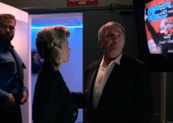 Damon Killian next to a television with joke credits on screen in The Running Man