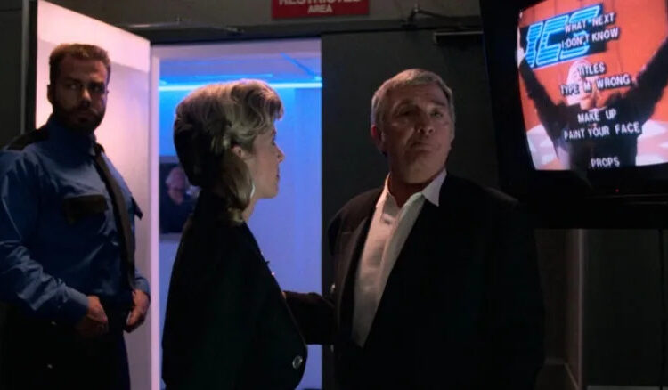 Damon Killian next to a television with joke credits on screen in The Running Man