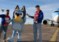 Bluey standing beside two men. They are wearing jeans, blue hoodies and Wales scarves and are holding Welsh flags. All three stand in front of the aeroplane.