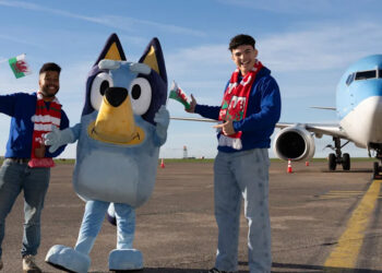 Bluey standing beside two men. They are wearing jeans, blue hoodies and Wales scarves and are holding Welsh flags. All three stand in front of the aeroplane.