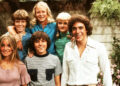 The cast of the Brady Bunch kids smiling outside the iconic house.