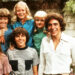 The cast of the Brady Bunch kids smiling outside the iconic house.