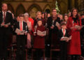 From left to right, William, George, Charlotte, Louis and Catherine in Westminster Abbey. Males are wearing black suits, white shirts; William a patterned dark red tie and both boys a red tie. Charlotte is wearing a dark red dress coat and Catherine is wearing a red dress coat with a large black bow at the neck. They are all holding lit candles and carol books and are standing. Behind them are people at the other pews.