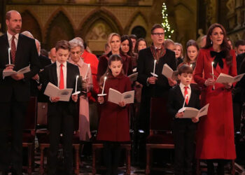 From left to right, William, George, Charlotte, Louis and Catherine in Westminster Abbey. Males are wearing black suits, white shirts; William a patterned dark red tie and both boys a red tie. Charlotte is wearing a dark red dress coat and Catherine is wearing a red dress coat with a large black bow at the neck. They are all holding lit candles and carol books and are standing. Behind them are people at the other pews.