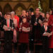 From left to right, William, George, Charlotte, Louis and Catherine in Westminster Abbey. Males are wearing black suits, white shirts; William a patterned dark red tie and both boys a red tie. Charlotte is wearing a dark red dress coat and Catherine is wearing a red dress coat with a large black bow at the neck. They are all holding lit candles and carol books and are standing. Behind them are people at the other pews.