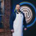Alfie Boe stood in front of the MasterChef logo smiling and wearing a white apron and every day clothes.