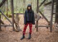 Claudia Winkleman stands in a forest wearing red tartan trousers, an oversized grey jumper and black boots.