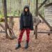 Claudia Winkleman stands in a forest wearing red tartan trousers, an oversized grey jumper and black boots.