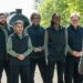 The finalists of The Celebrity Traitors stand in a group wearing matching black and green outfits, facing host Claudia Winkleman with a steam train in the background behind them.