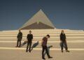 Four guys stand on steps in front of a pyramid-like building against a blue sky.