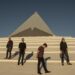 Four guys stand on steps in front of a pyramid-like building against a blue sky.