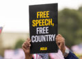 A demonstrator (with only their hands visible) holds up a sign that reads " FREE SPEECH, FREE COUNTRY - ACLU".