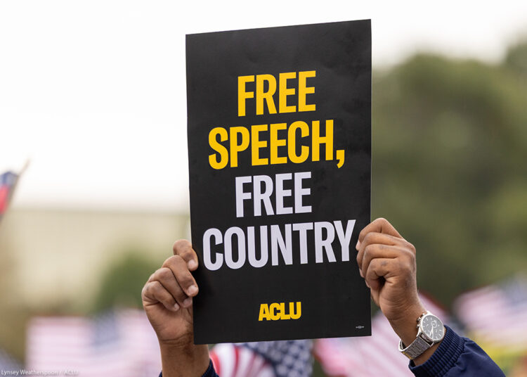 A demonstrator (with only their hands visible) holds up a sign that reads " FREE SPEECH, FREE COUNTRY - ACLU".
