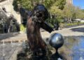 A statue and water fountain outside the Culver Theater in Downtown Culver City. A bi-pedal Bronze lion in a long coat poses on its legs. A silver sphere is propped up next to it. Little spouts of water surround them.