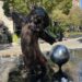 A statue and water fountain outside the Culver Theater in Downtown Culver City. A bi-pedal Bronze lion in a long coat poses on its legs. A silver sphere is propped up next to it. Little spouts of water surround them.