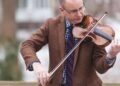 A white man wearing glasses and a brown sport coat plays a violin. Andy Carlson directs the Greenvillebillies, Bluegrass Ensemble concert