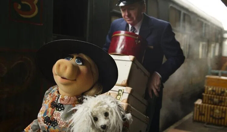 Miss Piggy carrying her dog while her butler carries her bags through a train station in Muppets Most Wanted