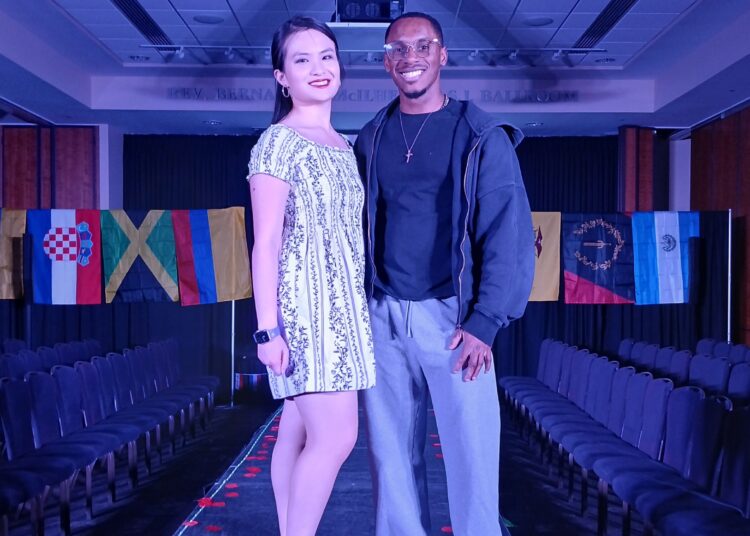 Two individuals pose on a fashion runway, with flags from various countries displayed in the background.